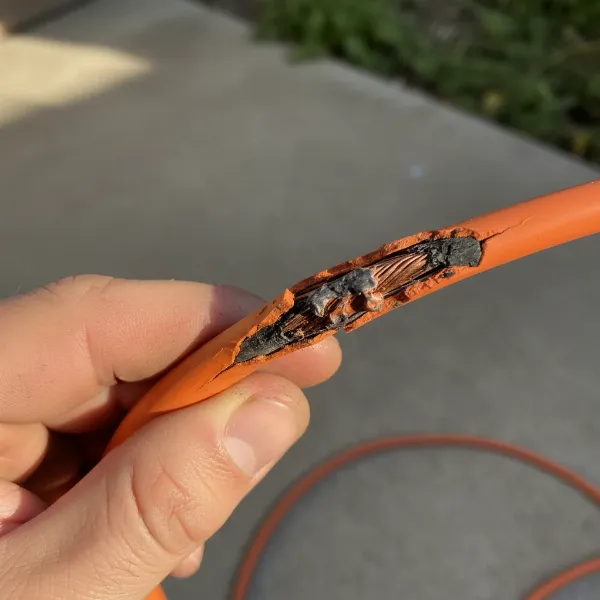 A close-up of a hand inspecting a worn or damaged section of an orange extension cord, showing frayed insulation or exposed wires, emphasizing the importance of regular safety checks.